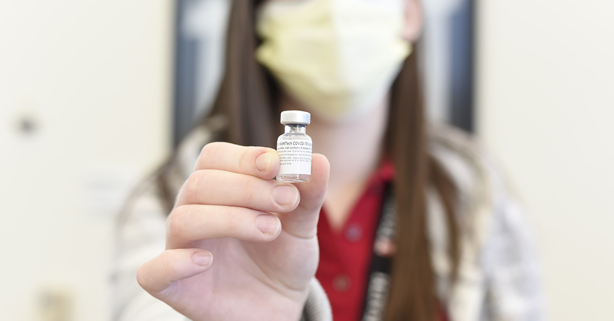 A photograph with a blurred background of a person holding a vial that reads "COVID-19 Vaccine".