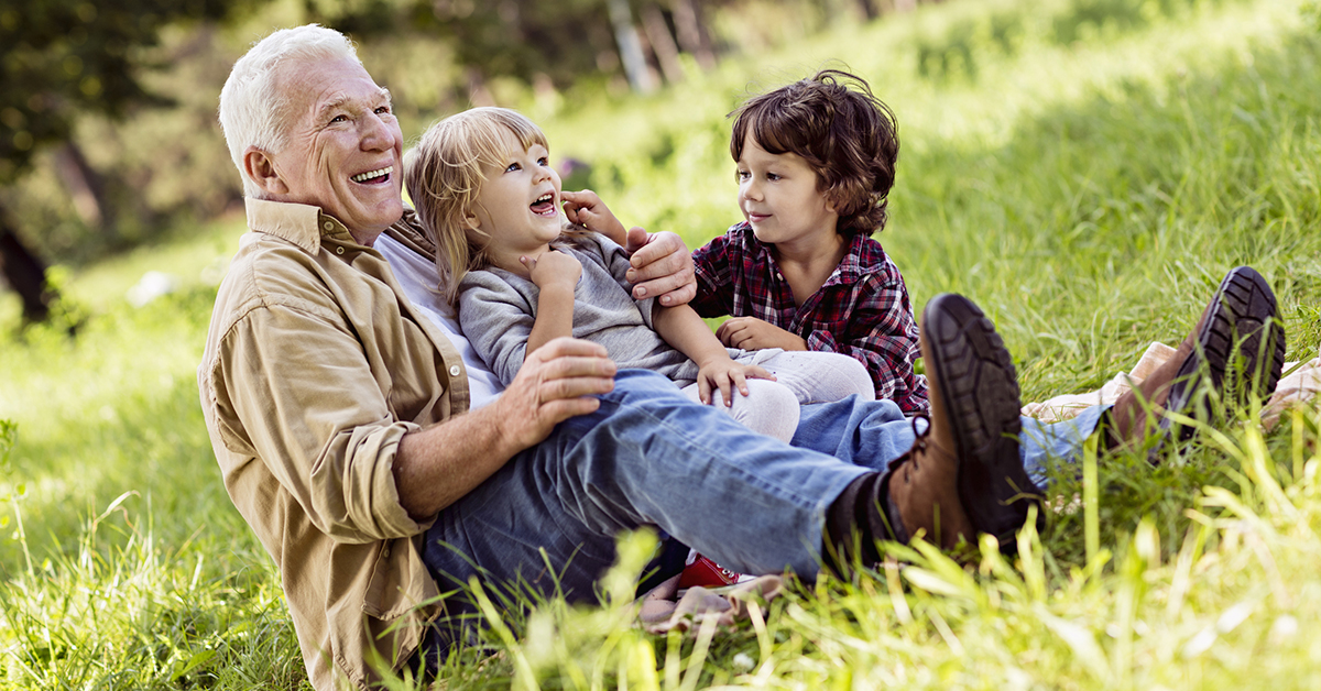 A photograph of an older man sitting in a grassy field with two children on his lap, all smiling widely.
