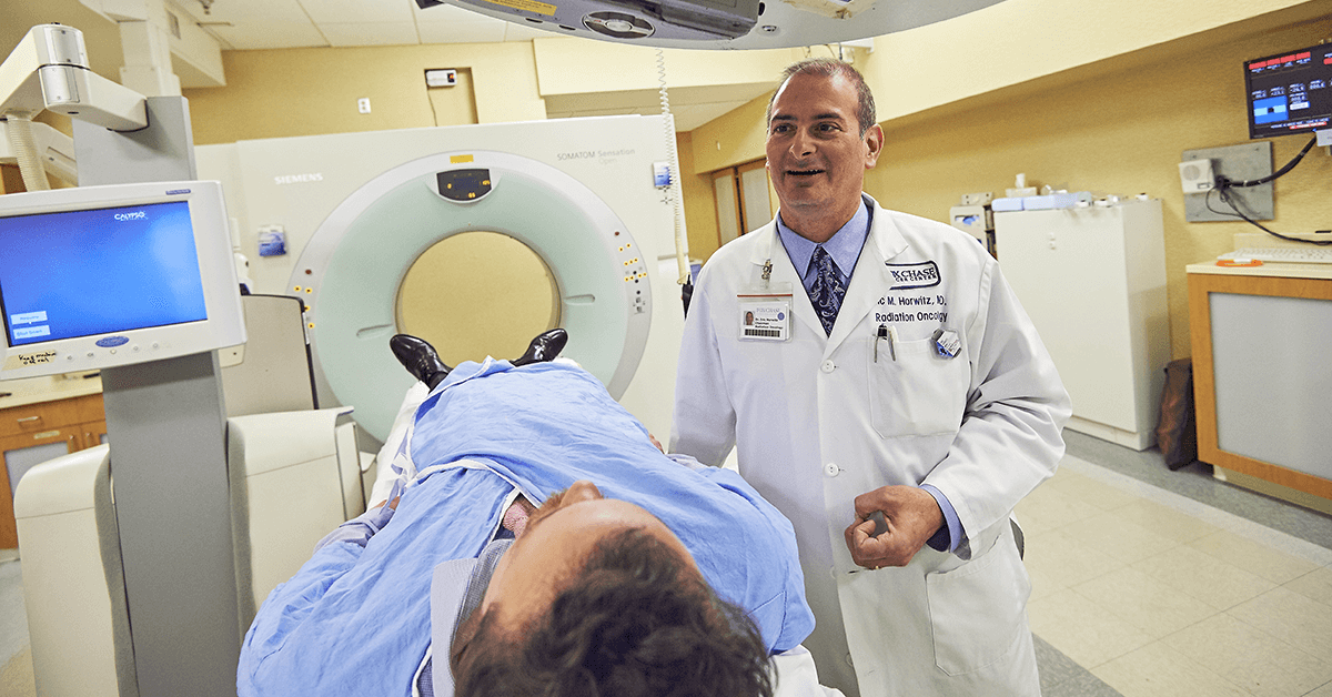 A photo of a Fox Chase doctor smiling as he stands next to a patient about to go into a medical imaging machine.
