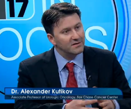 Dr. Alexander Kutikov on a TV show with the channel's logo in the background, speaking to someone off camera.
