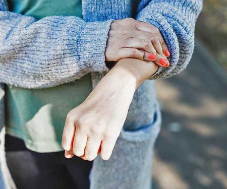 A closeup photo of a person's extended arm, which they touch with their other hand as they stand outside.