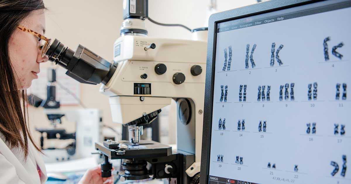 A photo of a Fox Chase doctor looking through a microscope, with a screen displaying chromosomes to their right.